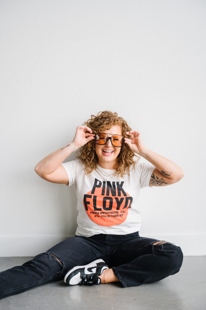 image of the author, redheaded female, sitting on the floor wearing distressed black jeans, sneakers, and a graphic t-shirt, holding sunglasses on her face. 