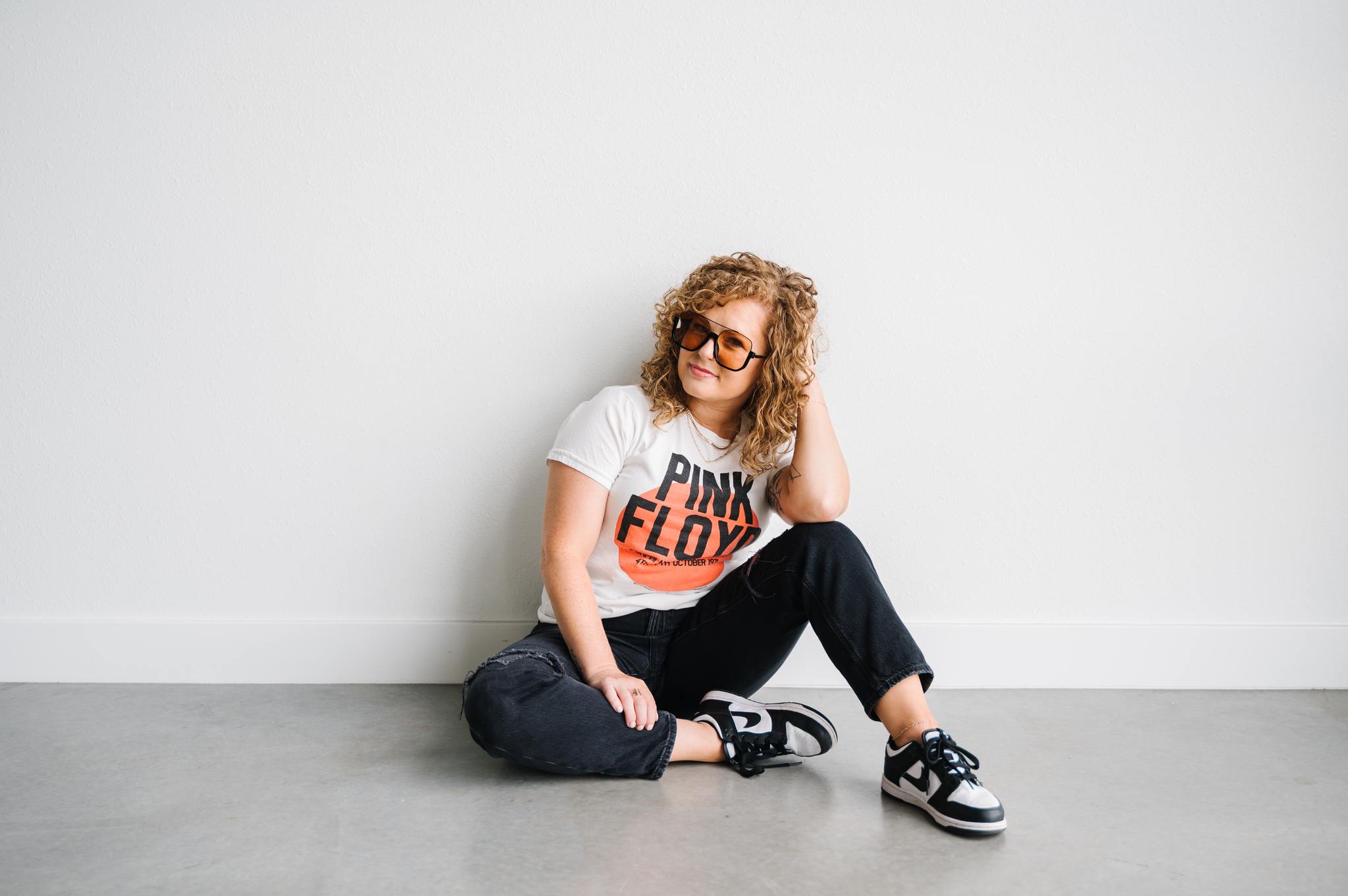 adult female with red curly hair wearing black jeans, graphic tee, and sneakers, sitting on ground, wearing sunglasses, hand in hair