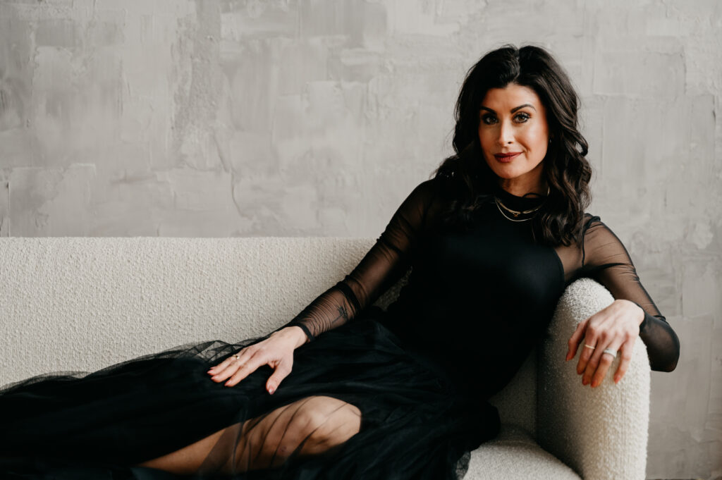brunette woman in black tulle dress, sitting on a white couch, looking at the camera