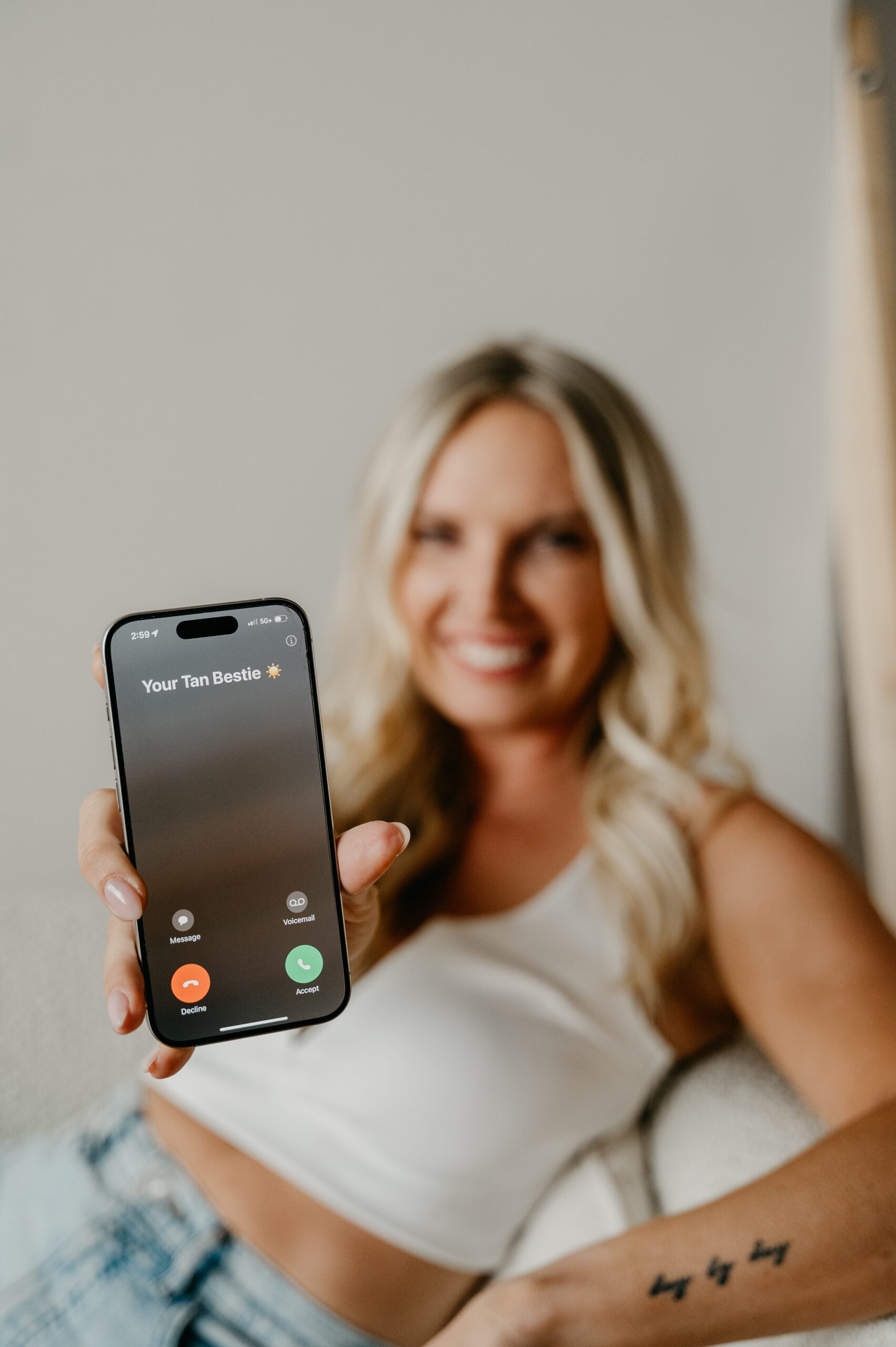 Blonde, smiling woman slightly blurred in background, holding out her phone, showing that "your tan bestie" is calling.